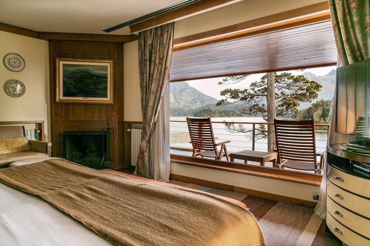 A bedroom with a bed, a fireplace, a painting, and a balcony with chairs and a view of mountains.