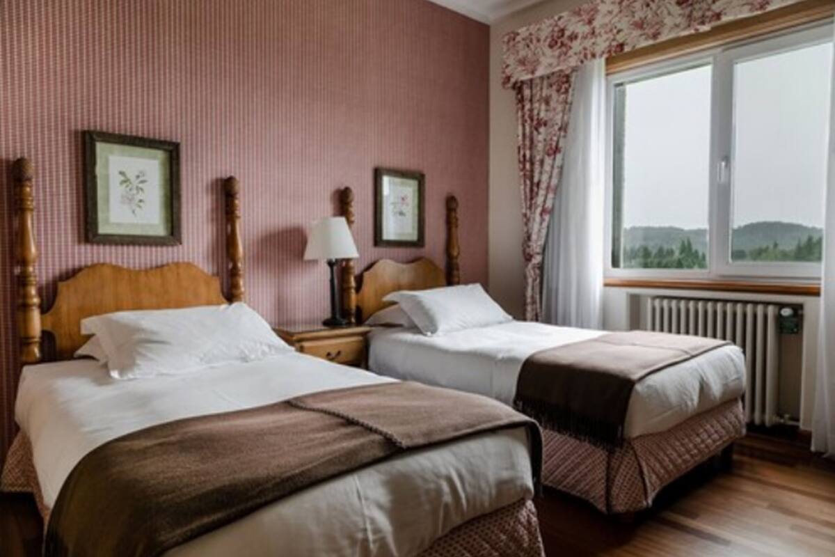 A hotel room with two beds, a radiator, a window with curtains, and framed pictures on the wall.