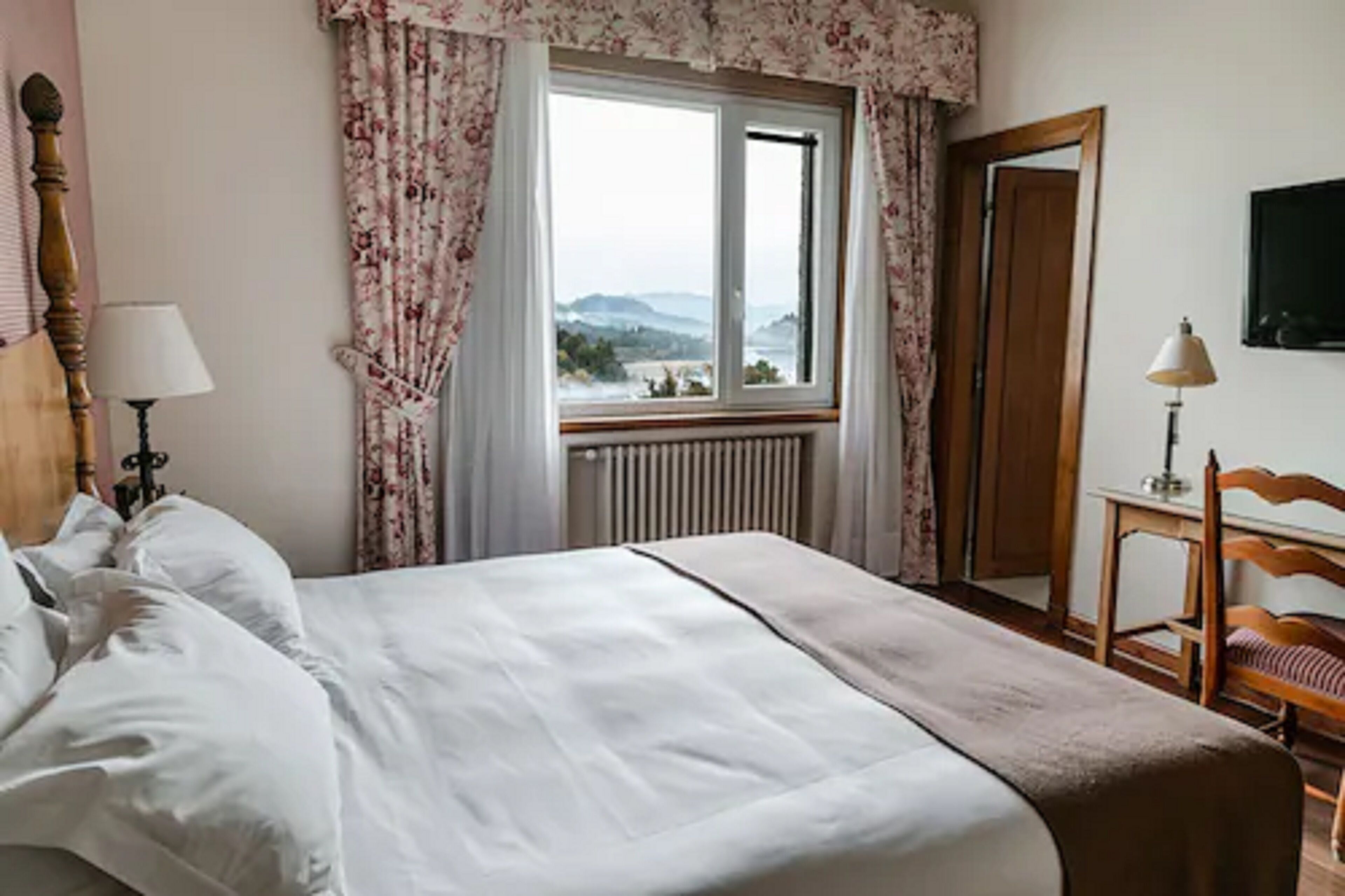 A bedroom with a bed, a window with curtains, a radiator, a wooden chair, and a mirror.