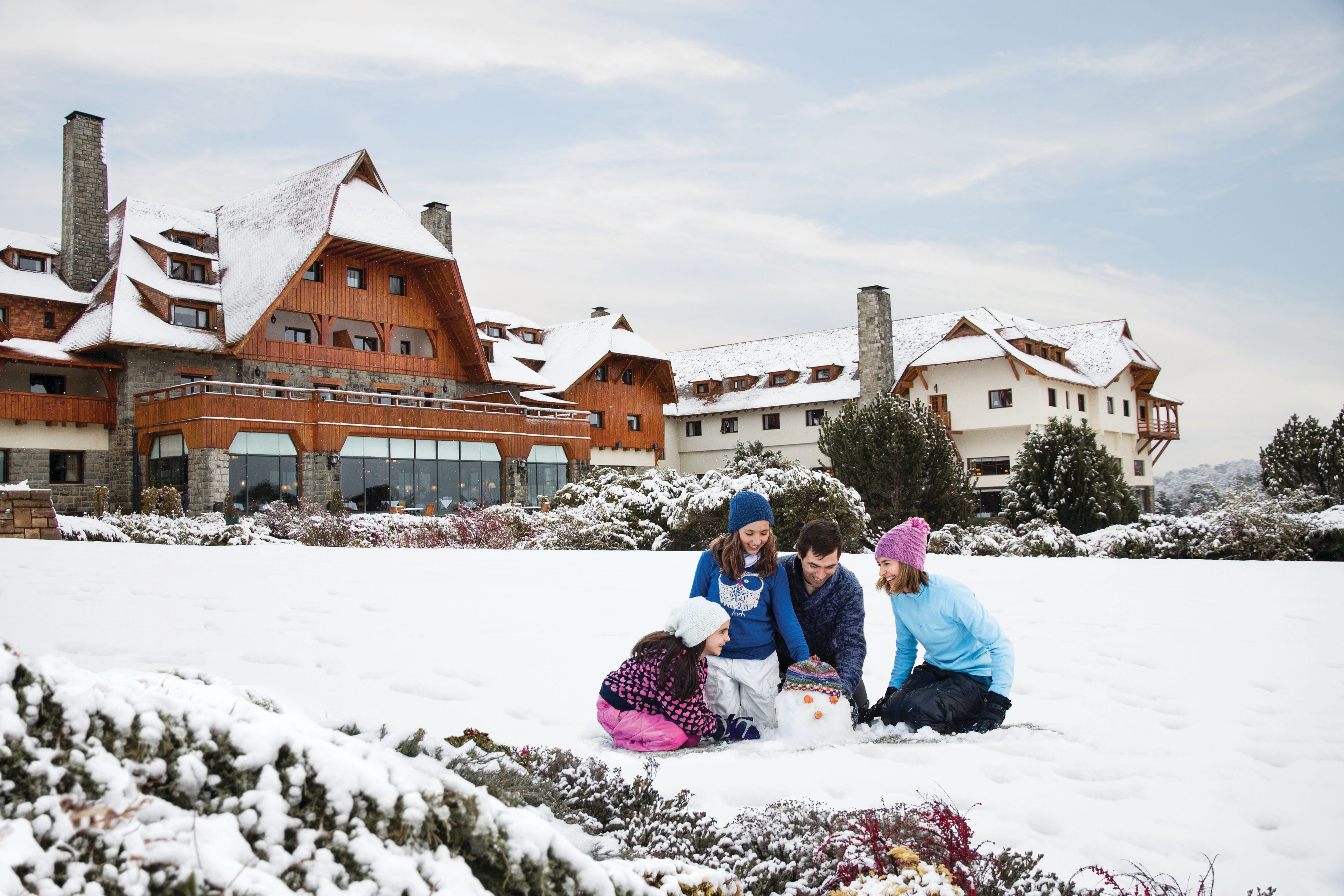 Eine Familie baut vor einem großen, rustikalen Gebäude mit vielen Fenstern und Balkonen einen Schneemann.