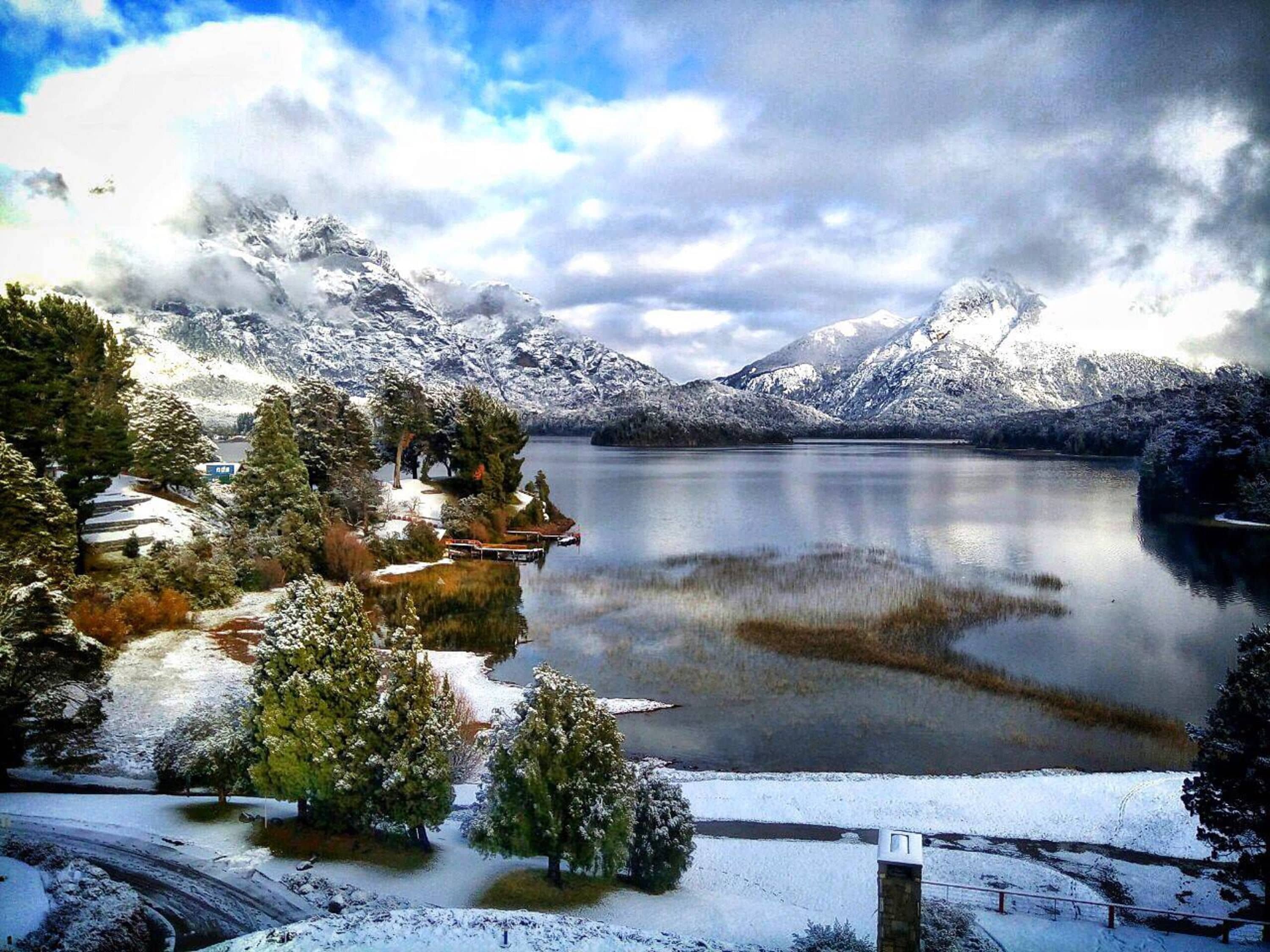 Un paisaje invernal sereno con un lago, montañas nevadas y un pequeño edificio con un muelle.