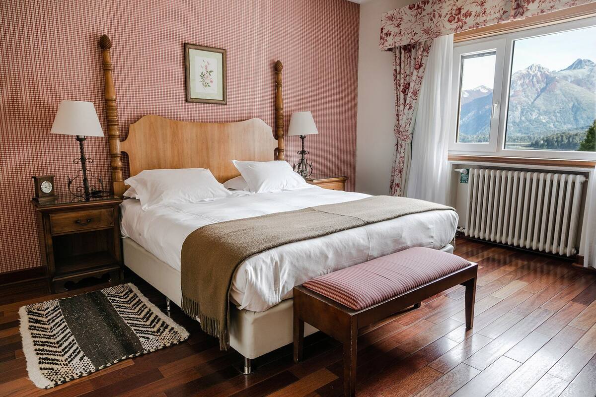 A neatly arranged bedroom with a large bed, two bedside tables with lamps, a bench, a radiator, and a window with a mountain view.