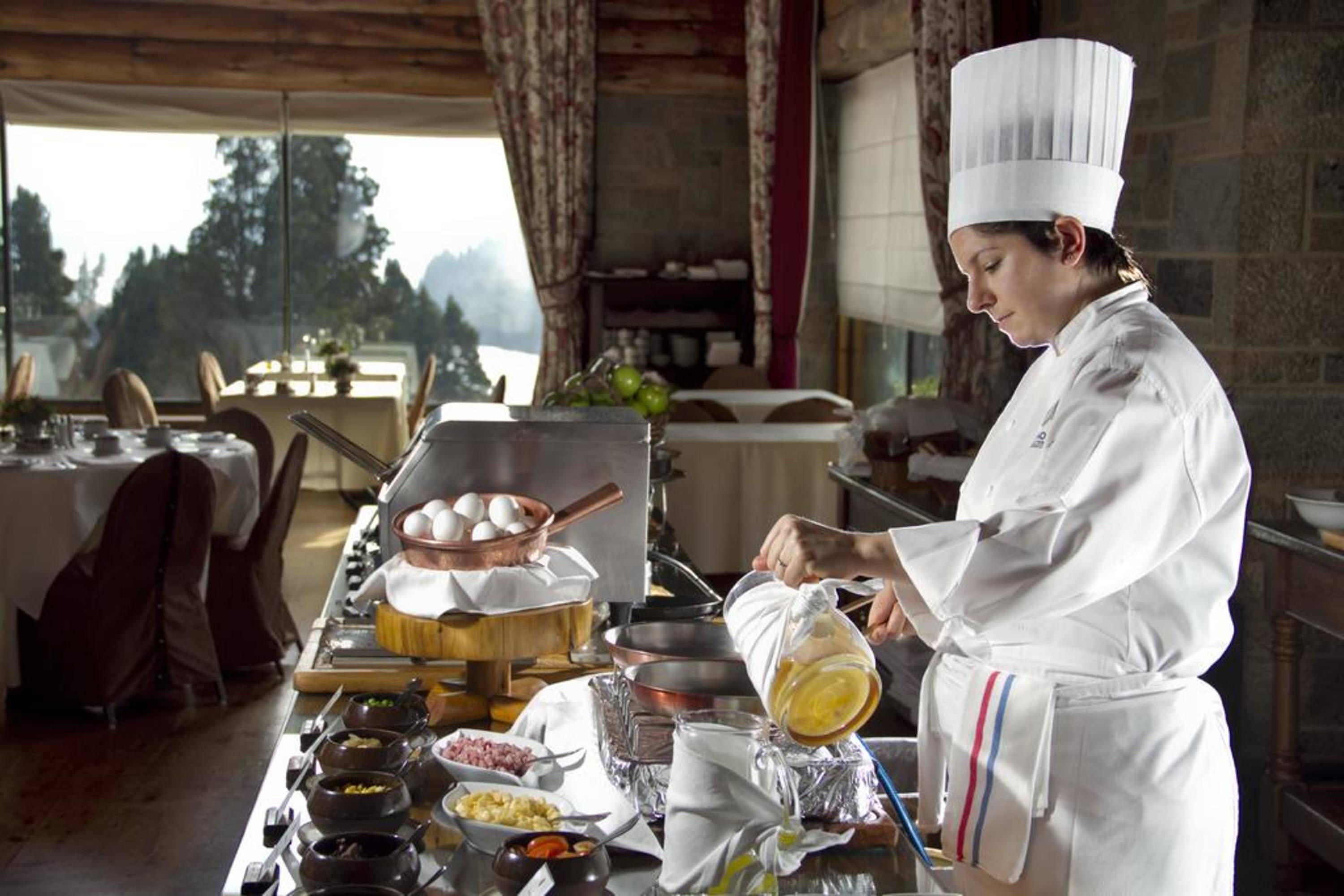 Un chef con uniforme blanco preparando comida en una cocina rústica con elementos de madera y vistas a árboles.