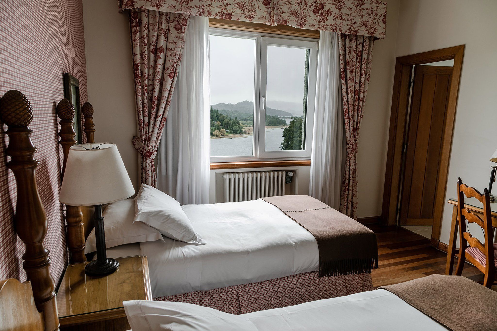 A bedroom with two beds, a wooden floor, a window with a view of a river and hills, and a door to the right.