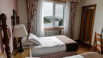 A bedroom with two beds, a wooden floor, a window with a view of a river and hills, and a door to the right.