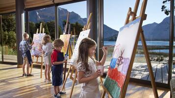 A group of children painting in a room with large windows overlooking a lake and mountains.