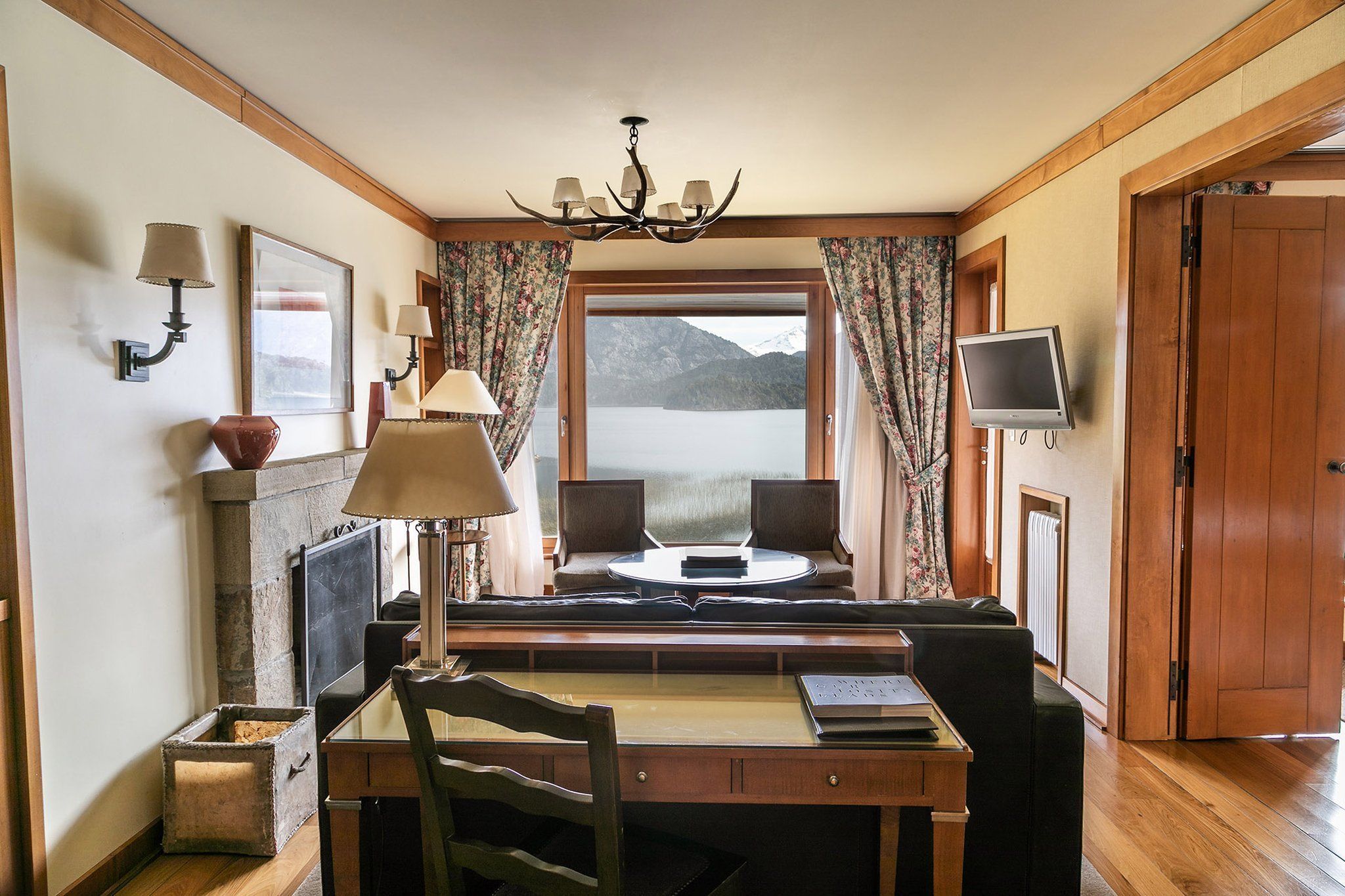 Een gezellige kamer met een houten bureau, een comfortabele stoel, een open haard, een televisie en een groot raam met uitzicht op een schilderachtig berglandschap.