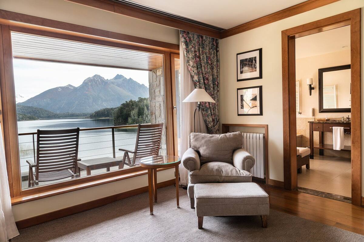 A room with a large window overlooking a lake and mountains, a sofa, a chair, a lamp, and a bathroom visible through an open door.