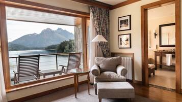 Una habitación con un ventanal grande que da a un lago y montañas, un sofá, una silla, una lámpara y un baño visible a través de una puerta abierta.
