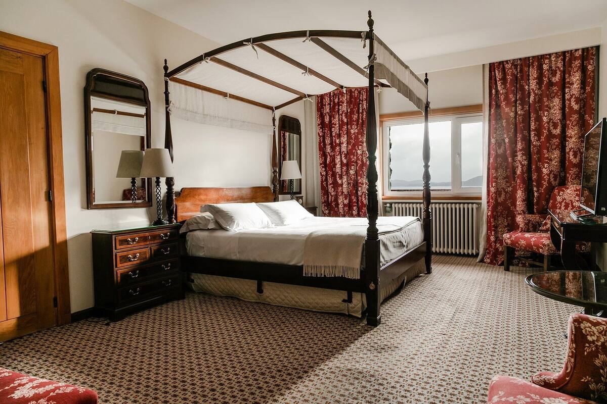 A four-poster bed with a canopy, a wooden dresser, a red upholstered chair, and a view of the outdoors through a window.
