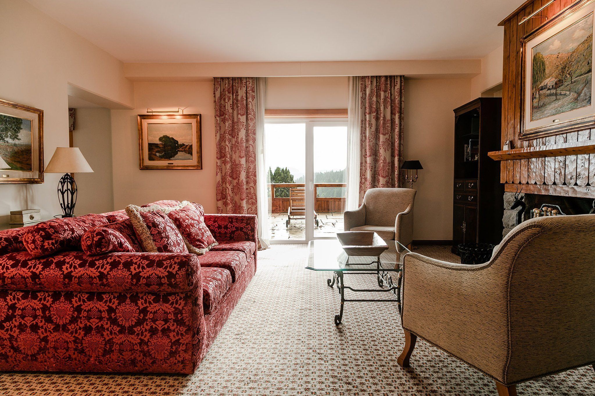 Una sala de estar con un sofá rojo estampado, un sillón color beige, una mesa de centro de vidrio y una chimenea. Hay un ventanal con cortinas, un cuadro en la pared y vistas a un balcón.