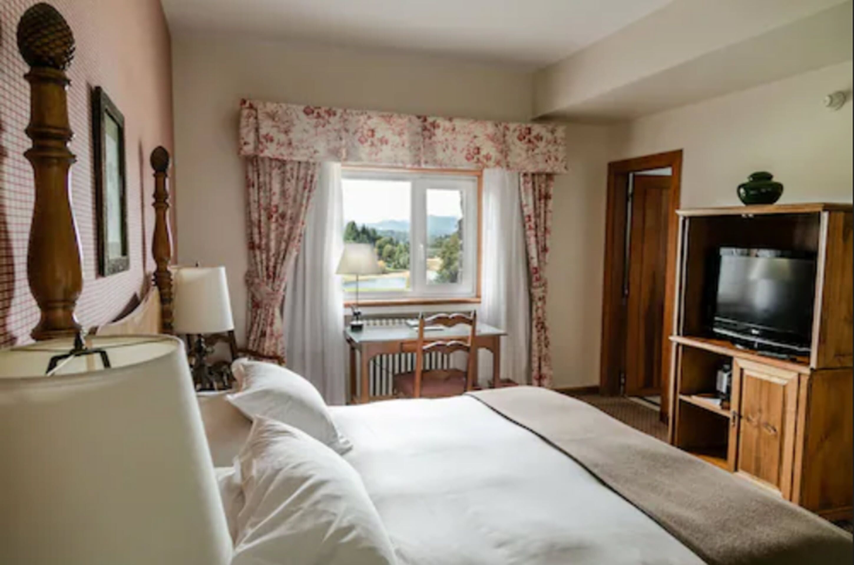 A hotel room with a bed, a desk with a chair, a television, and a window with curtains.