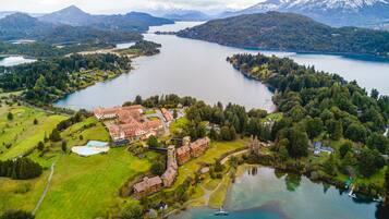 An aerial view of a lakeside property with a large building, possibly a hotel, surrounded by lush greenery and a mountain in the background.