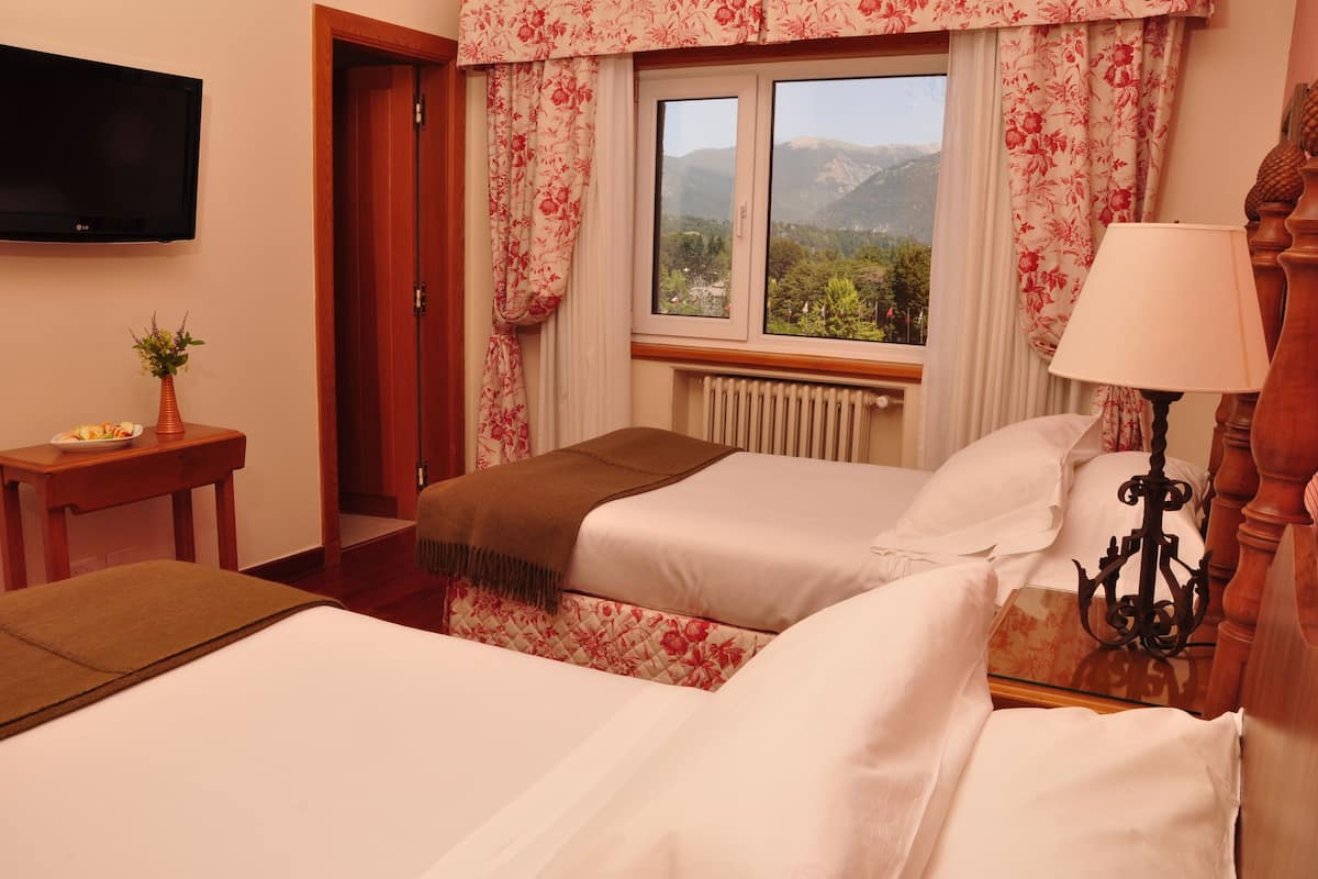 A hotel room with two beds, a TV, a window with a mountain view, a bedside table, and a lamp.