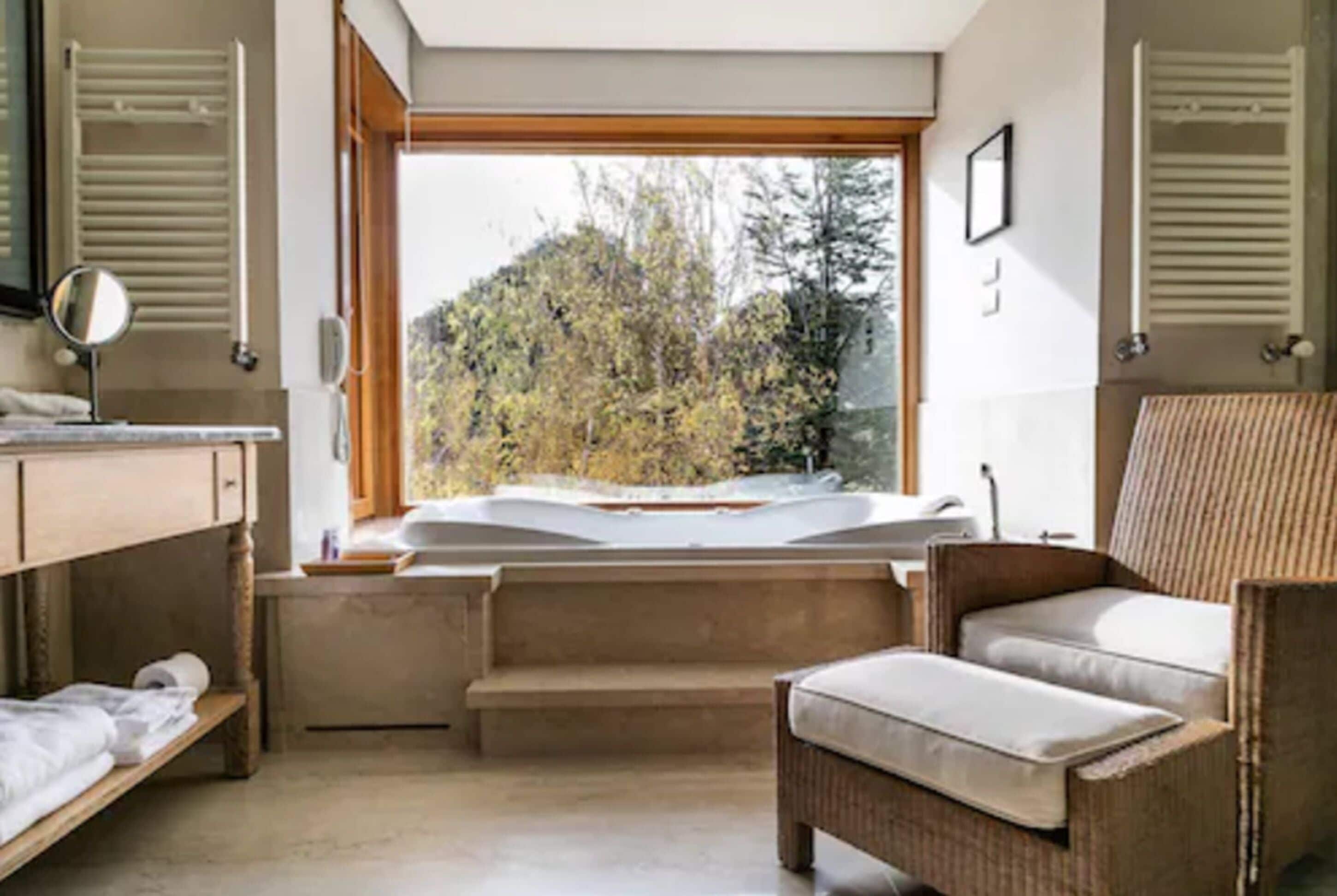 Un dormitorio con cama, una silla, un ventanal con vistas a árboles y una mesita con espejo.