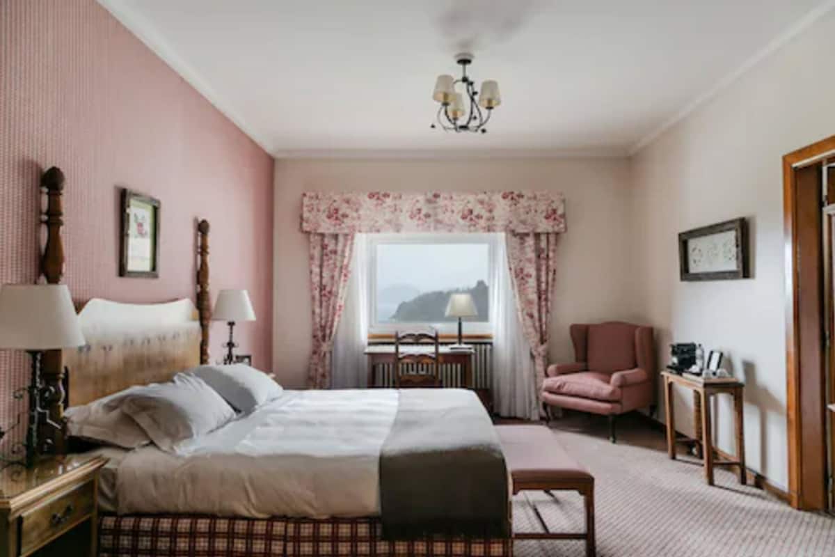 A bedroom with a bed, bedside tables, a chair, a window with curtains, and a chandelier.