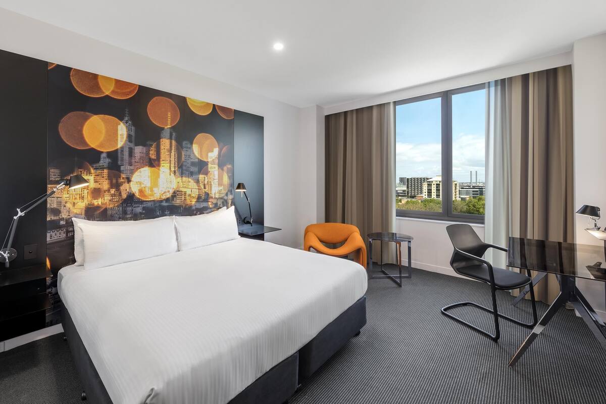 A hotel room with a large bed, a cityscape mural, a desk with a chair, and a view of the city through large windows.