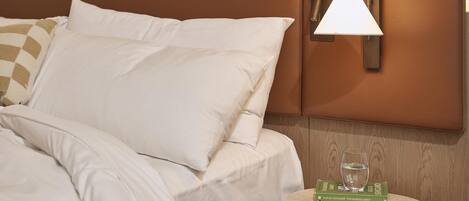 Un lit bien fait, avec du linge de lit blanc, une tête de lit marron, une lampe fixée au mur et une petite table ronde sur laquelle se trouvent un livre et un verre d’eau.