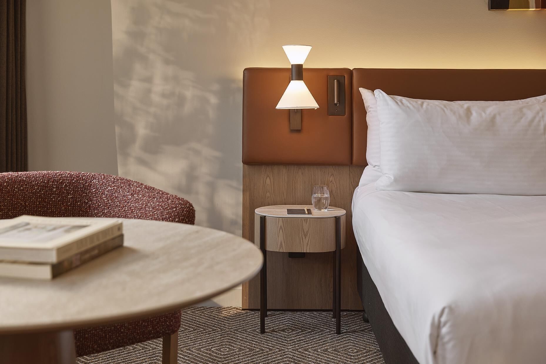 Una camera d'albergo con un letto, un comodino con una lampada, un tavolino rotondo con un libro e una sedia.