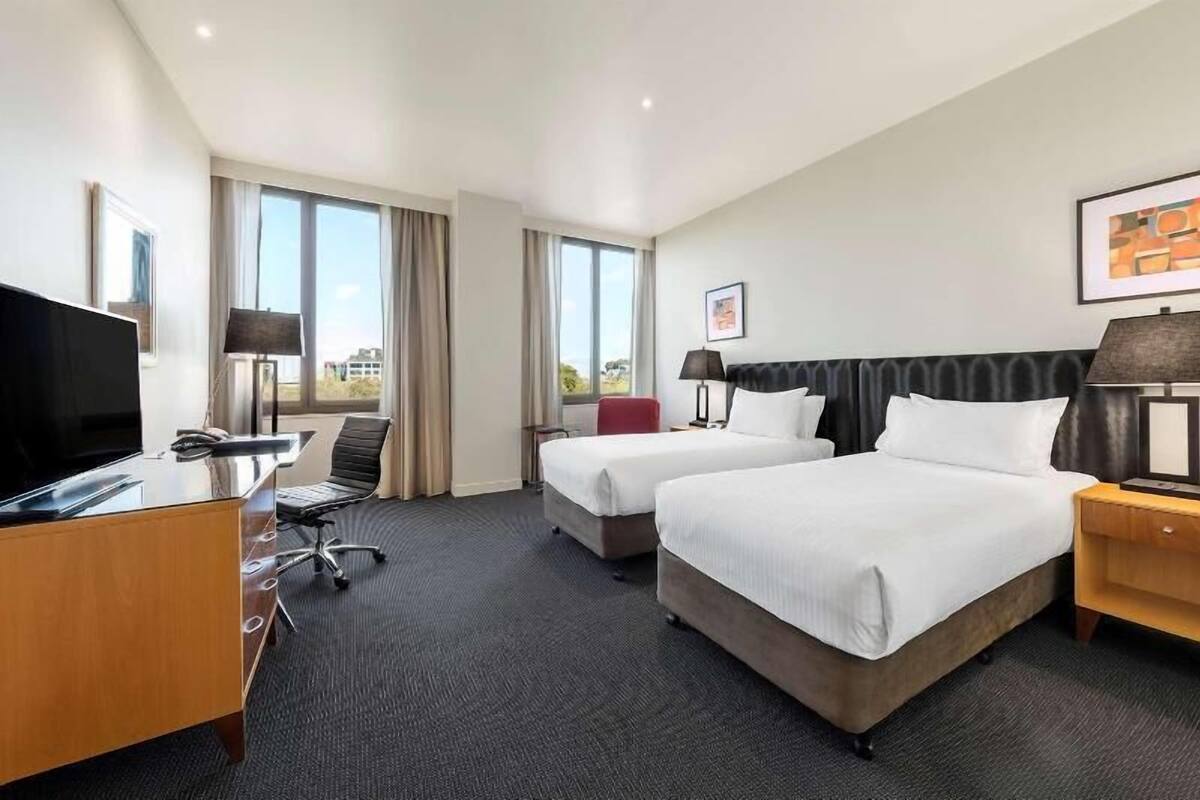 A hotel room with two beds, a desk, a TV, and a window with curtains.