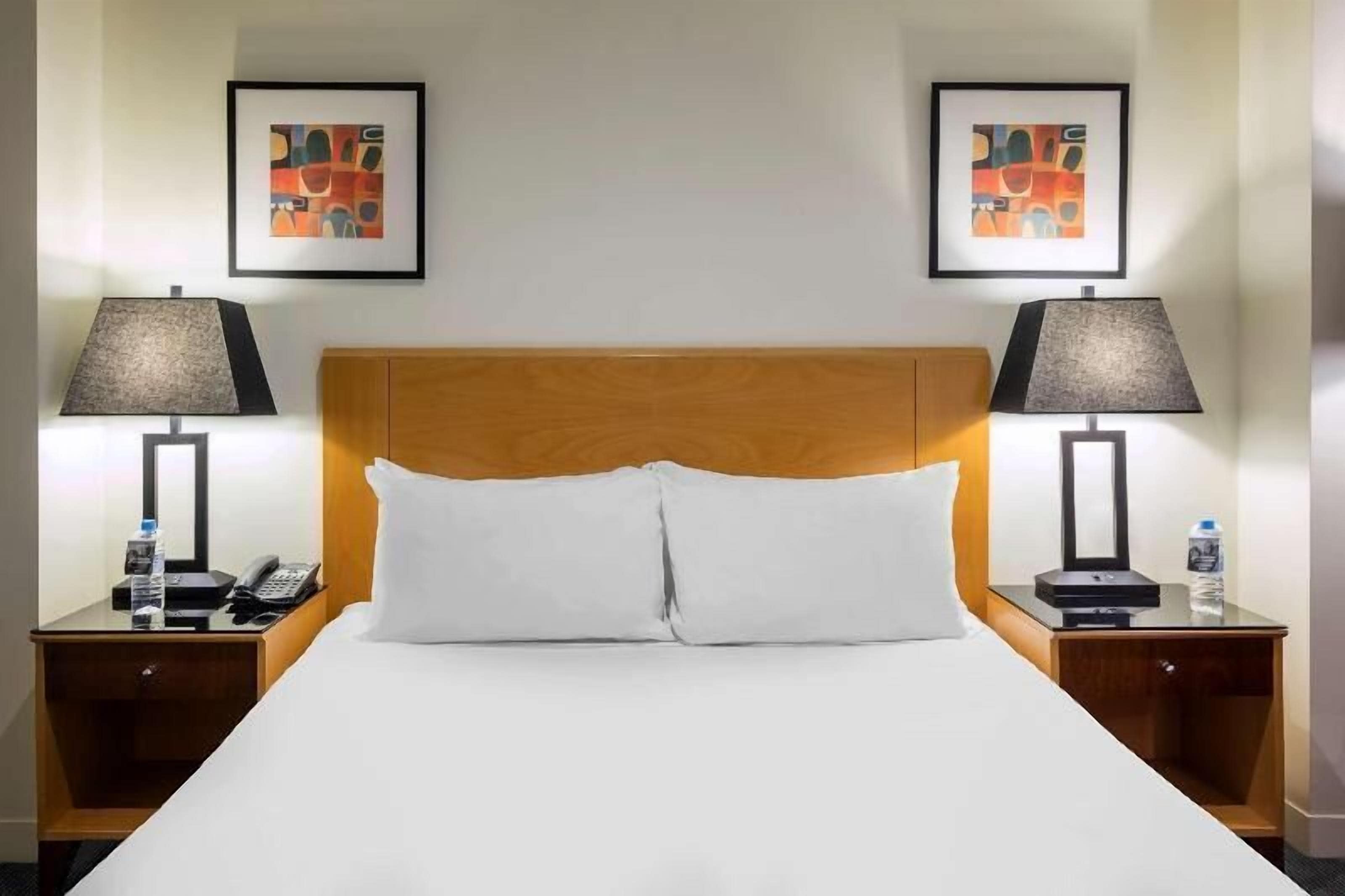 A hotel room with a large bed, two nightstands, two lamps, a phone, a water bottle, and two framed abstract paintings on the wall.