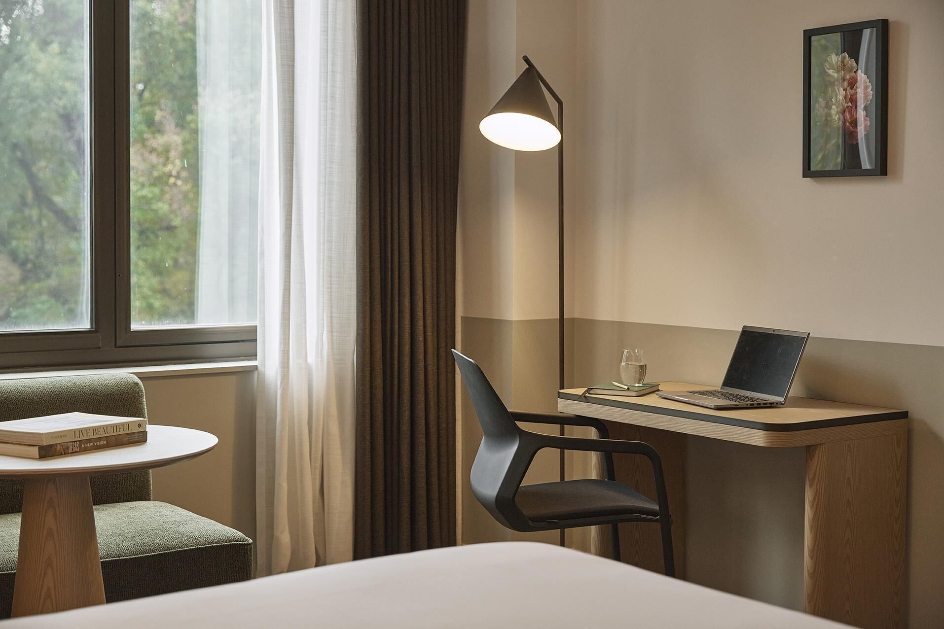 A hotel room with a bed, a desk with a laptop, a chair, a round table with books, a floor lamp, and a window with curtains.