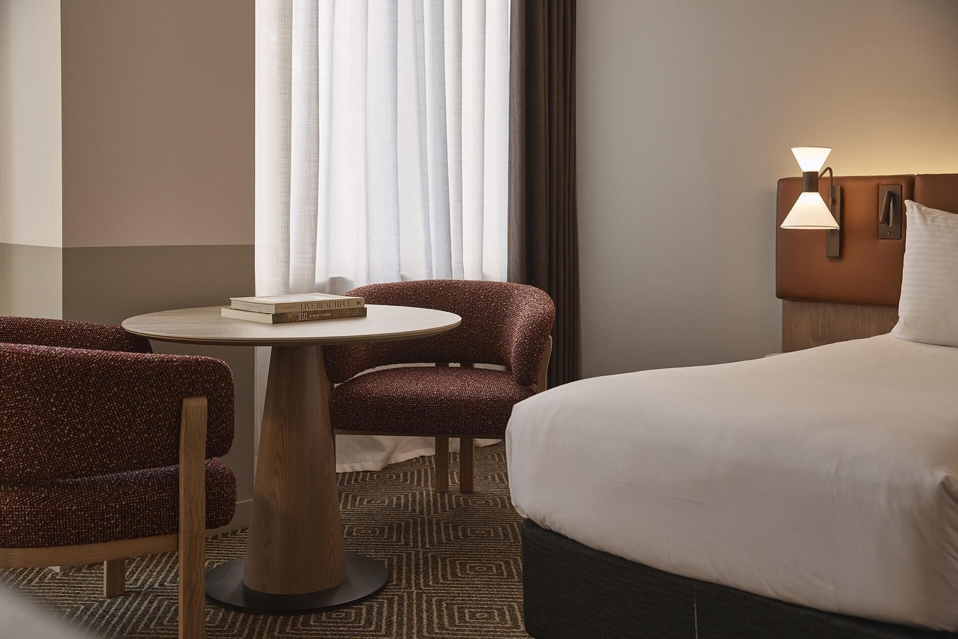 Ett hotellrum med en säng, två stolar, ett litet runt bord, en lampa och ett fönster med gardiner.