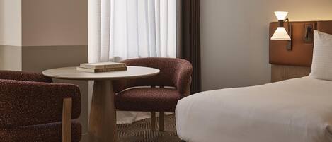 A hotel room with a bed, two chairs, a small round table, a lamp, and a window with curtains.