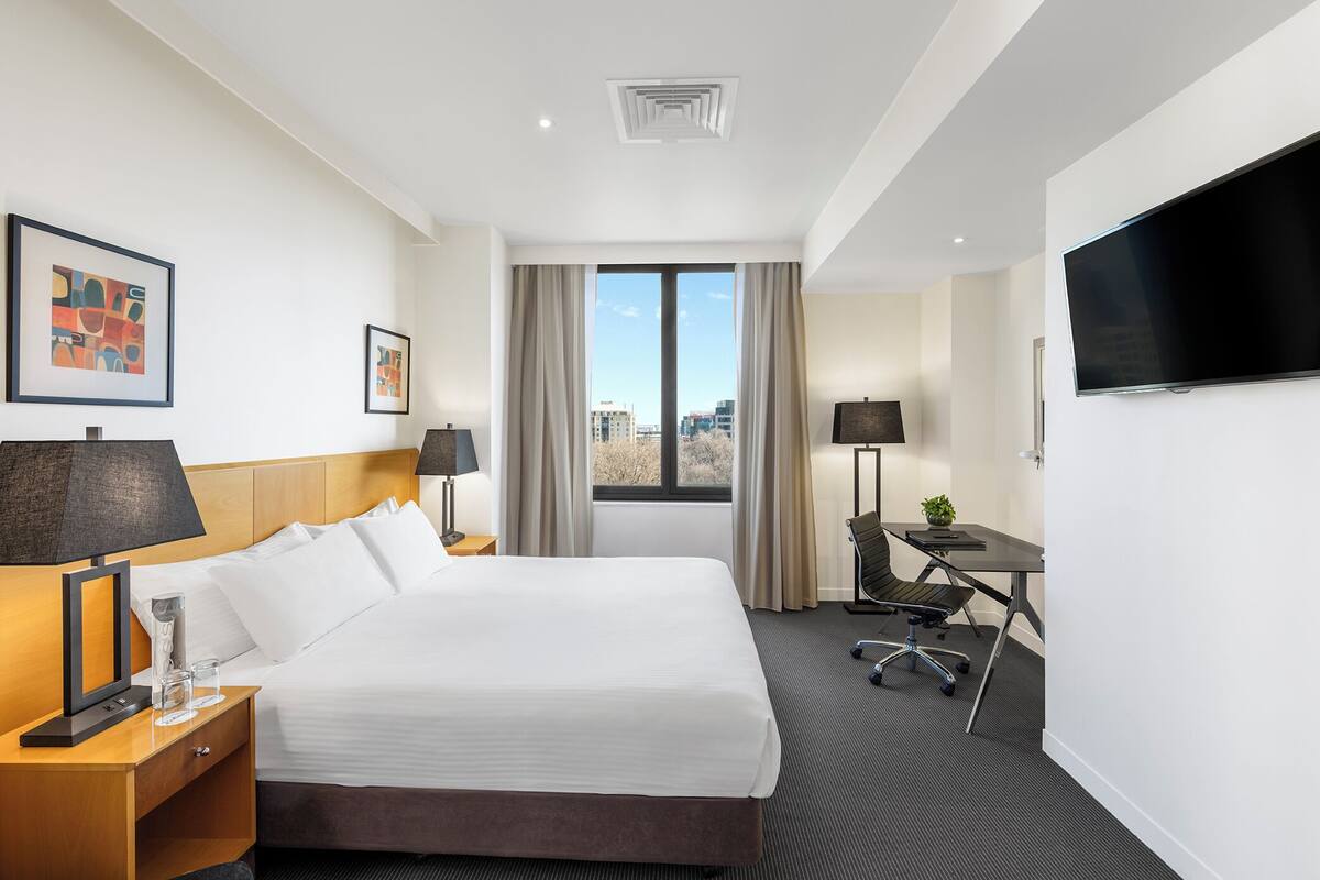 A hotel room with a large bed, a desk with a chair, a flat-screen TV, and a window with a city view.