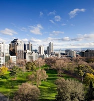 Paisagem urbana com edifícios modernos altos, um parque com grama verde e árvores, e um céu azul claro.