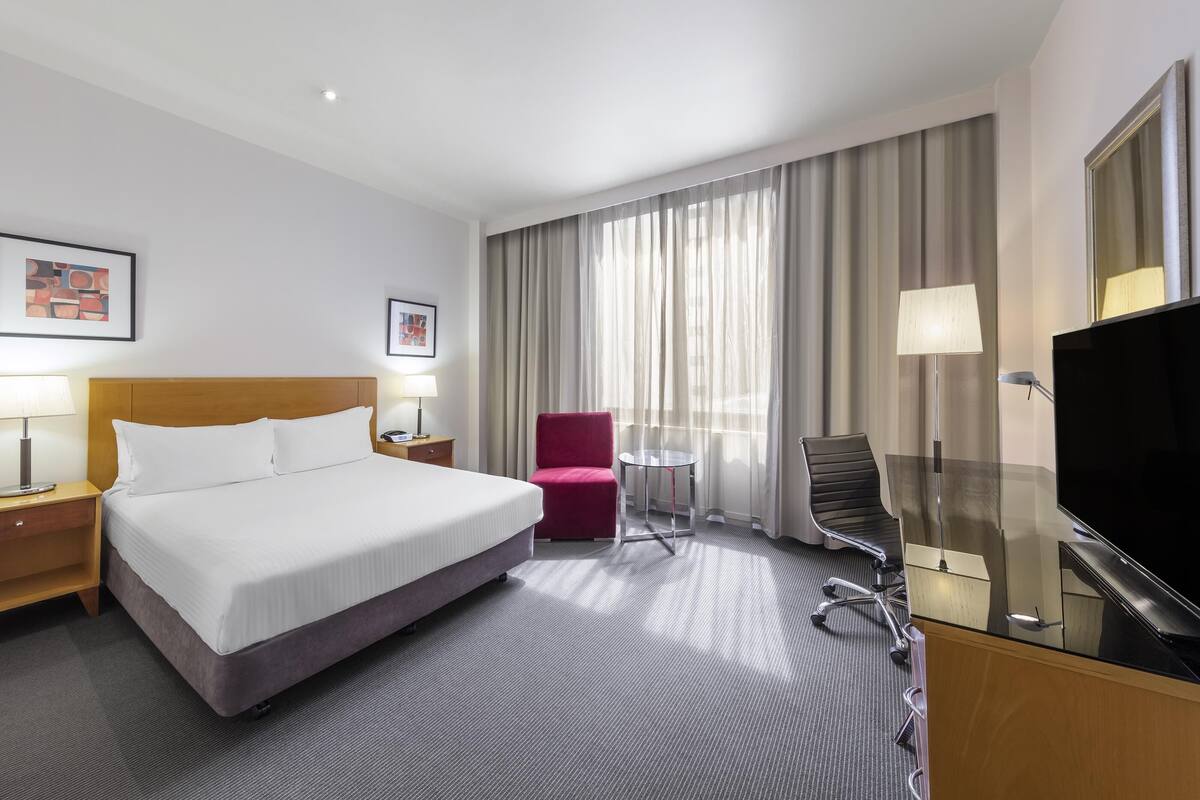 A hotel room with a bed, a desk with a TV, a chair, a small table, and curtains.