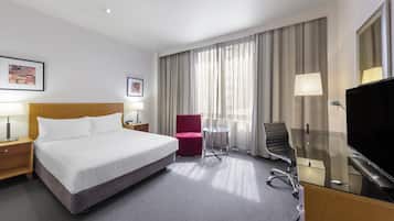 A hotel room with a bed, a desk with a TV, a chair, a small table, and curtains.