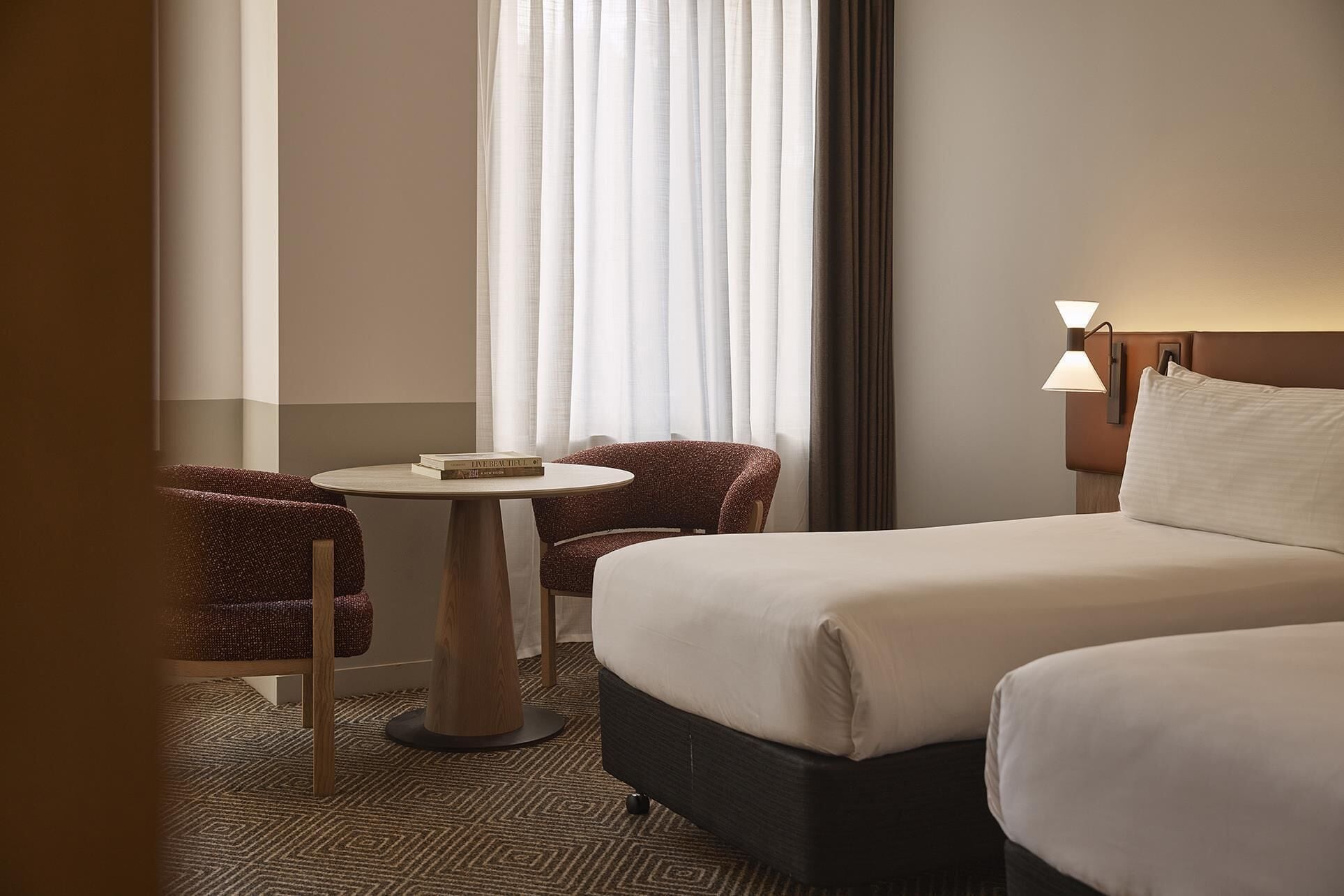 A hotel room with a bed, two chairs, a small table, a lamp, and a window with curtains.