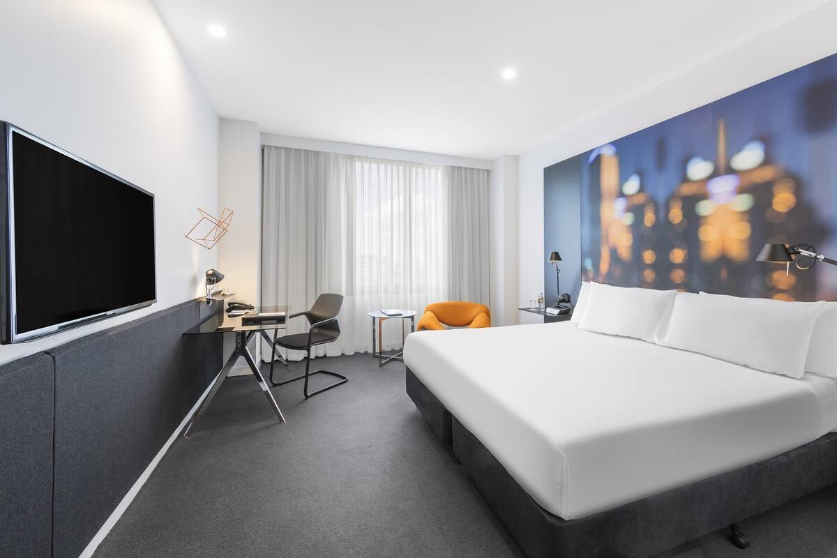 A hotel room with a large bed, a desk with a chair, a TV, and a cityscape painting on the wall.