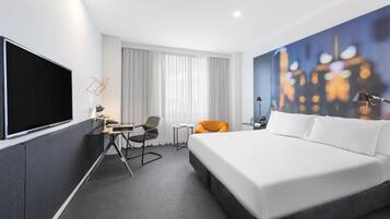 A hotel room with a large bed, a desk with a chair, a TV, and a cityscape painting on the wall.
