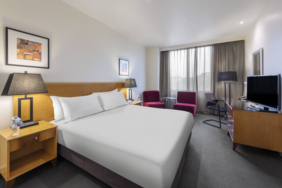 A hotel room with a large bed, a desk with a TV, a chair, a small table, and a lamp.