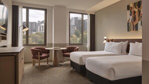A hotel room with two beds, a desk, a chair, a small table, and large windows offering a city view. - Flagstaff Gardens Hotel Melbourne (Melbourne)