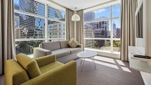 Une chambre d’hôtel moderne dotée d’une grande fenêtre donnant sur la ville, d’un canapé gris, d’un fauteuil vert et d’une petite table blanche.