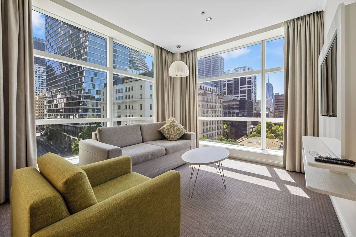 Une chambre d’hôtel moderne dotée d’une grande fenêtre donnant sur la ville, d’un canapé gris, d’un fauteuil vert et d’une petite table blanche.