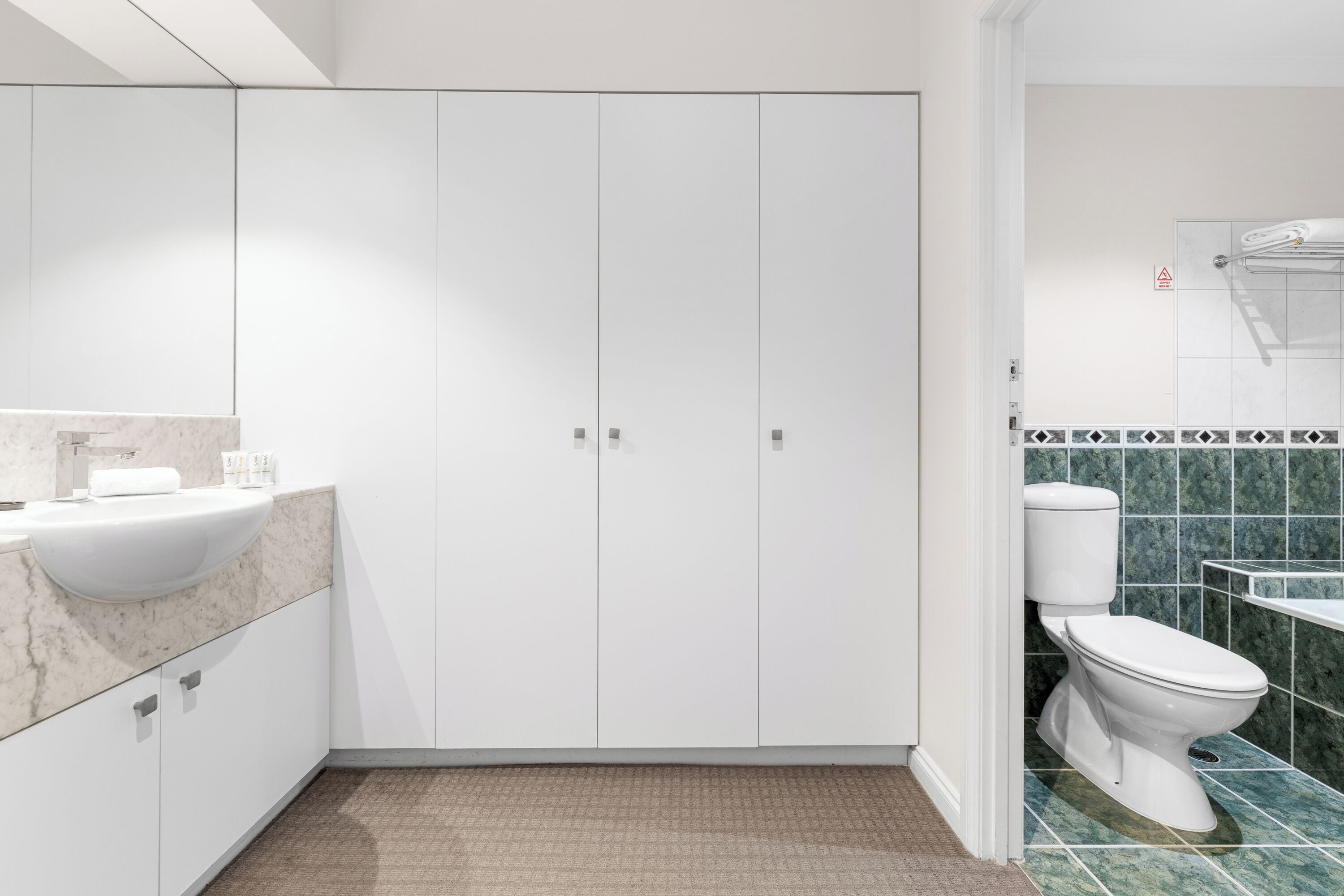 Une salle de bain moderne avec un meuble-lavabo blanc, un grand miroir et des toilettes dont le mur est recouvert de carreaux verts.