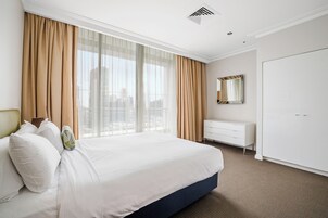 Habitación de hotel con una cama grande, un tocador blanco, un espejo y vistas de la ciudad a través de cortinas translúcidas.