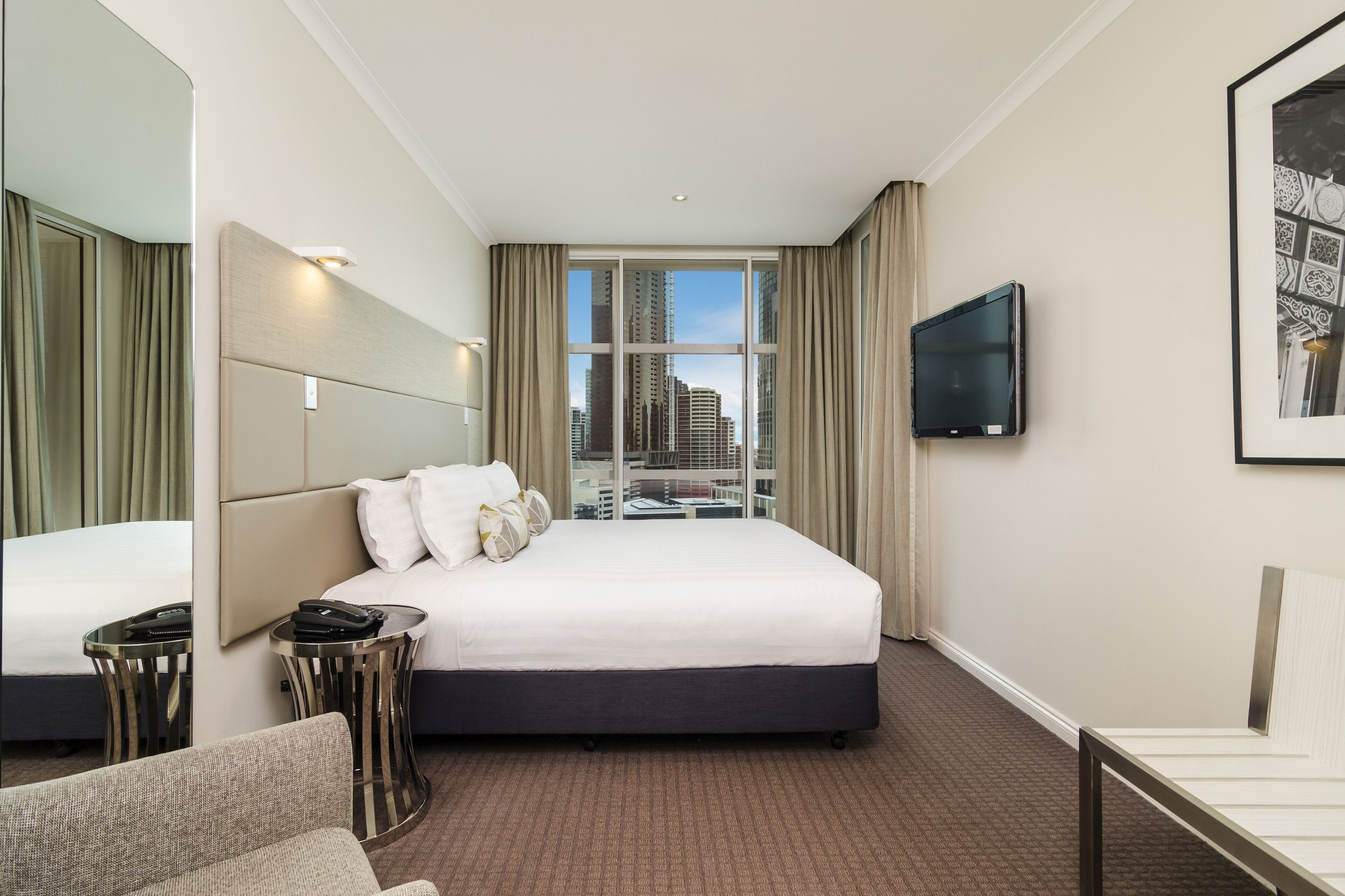 Ein Hotelzimmer mit einem großen Bett, einem Fernseher, Blick auf die Stadt und einem Spiegel.
