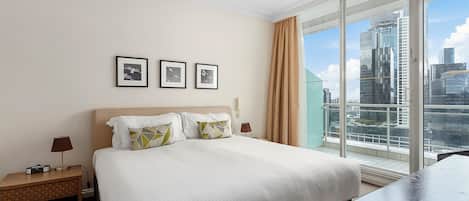 Habitación de hotel con una cama grande, una mesita de noche con lámpara y vistas a un paisaje urbano a través de amplios ventanales.