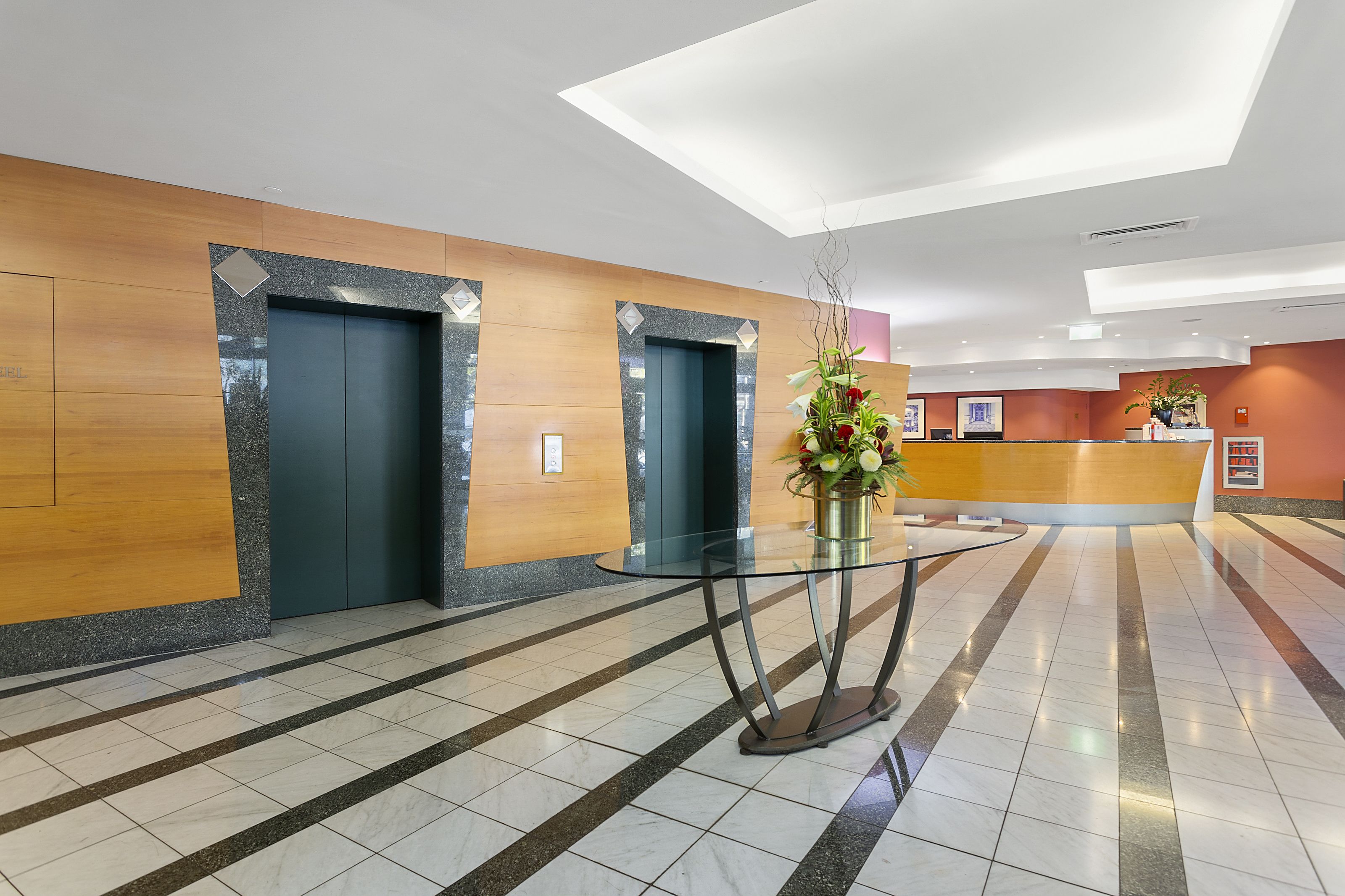 A modern lobby with a reception desk, two elevators, and a glass table with a vase of flowers.
