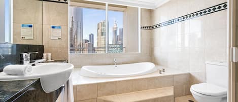 A modern bathroom with a large bathtub, two sinks, and a toilet. A cityscape is visible through the window.