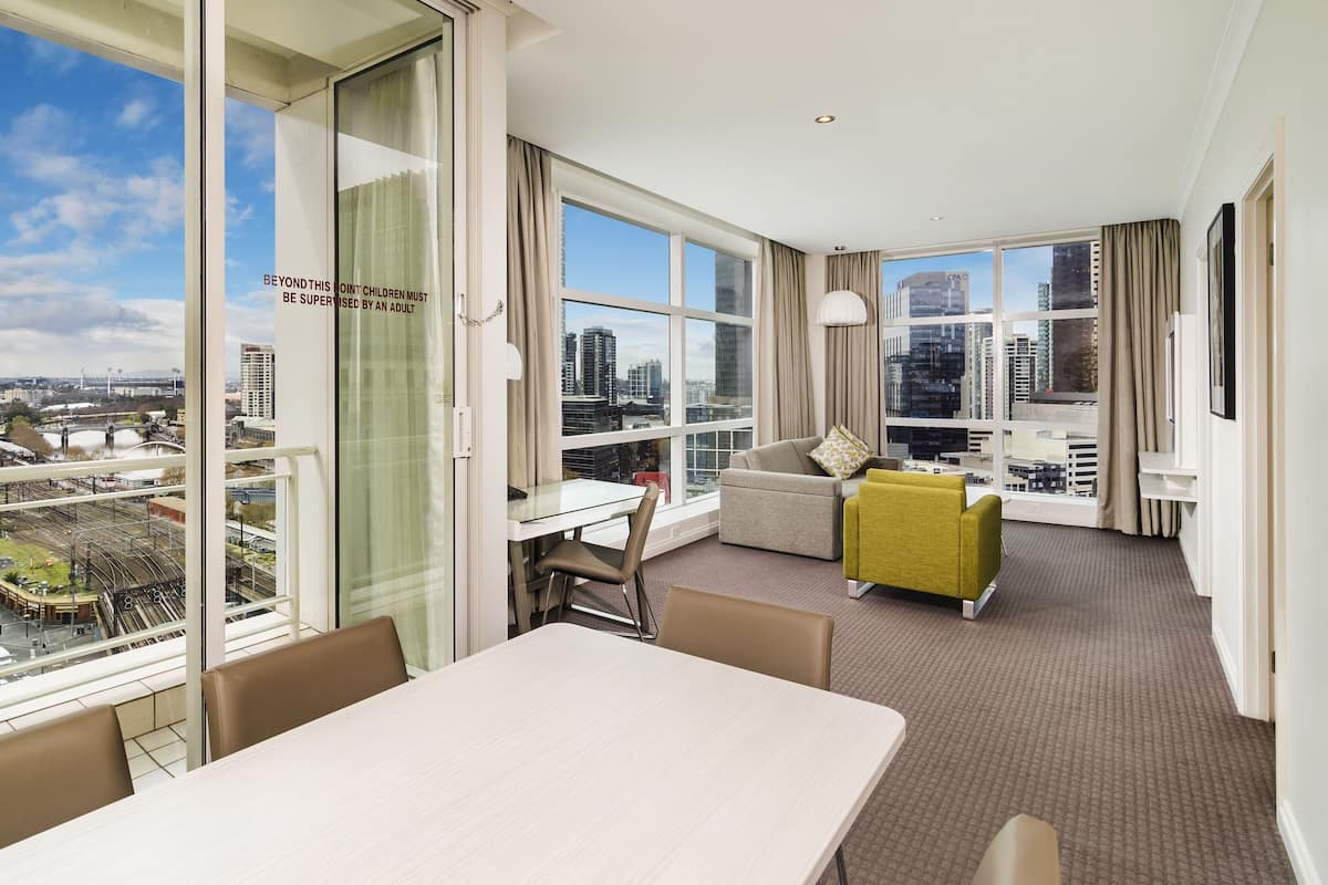 Une chambre d’hôtel moderne dotée d’une grande fenêtre donnant sur la ville, d’un coin repas et d’un espace salon avec un canapé et des fauteuils.