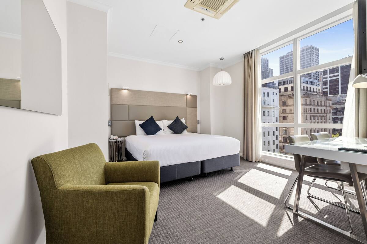 Une chambre d’hôtel avec un lit, un fauteuil vert, un bureau et une grande fenêtre donnant sur la ville.