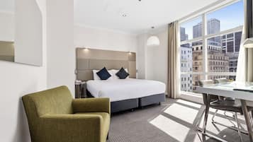 A hotel room with a bed, a green armchair, a desk, and a large window offering a city view.