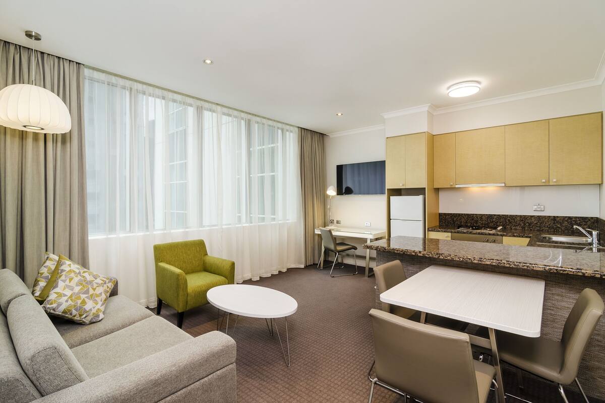 Une chambre d’hôtel comprenant un coin salon, une table à manger, une cuisine avec un évier et une grande fenêtre avec des rideaux.