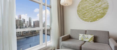 Una sala de estar moderna con un sofá gris, una mesa redonda y vistas de la ciudad a través de amplios ventanales.